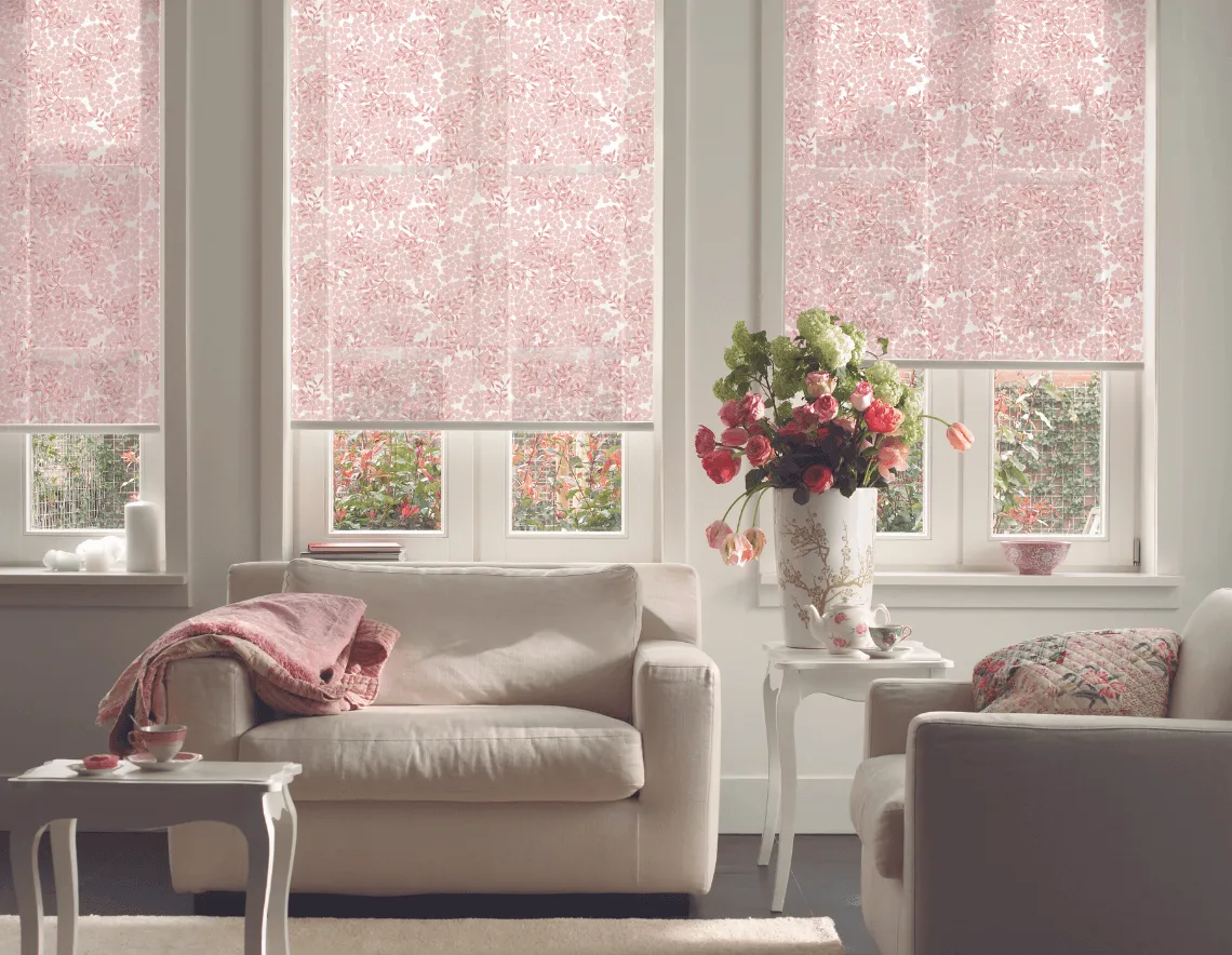 Cortinas Roller estampadas, en color rosa. Con muebles y paredes blancas y manta nórdica haciendo juego.