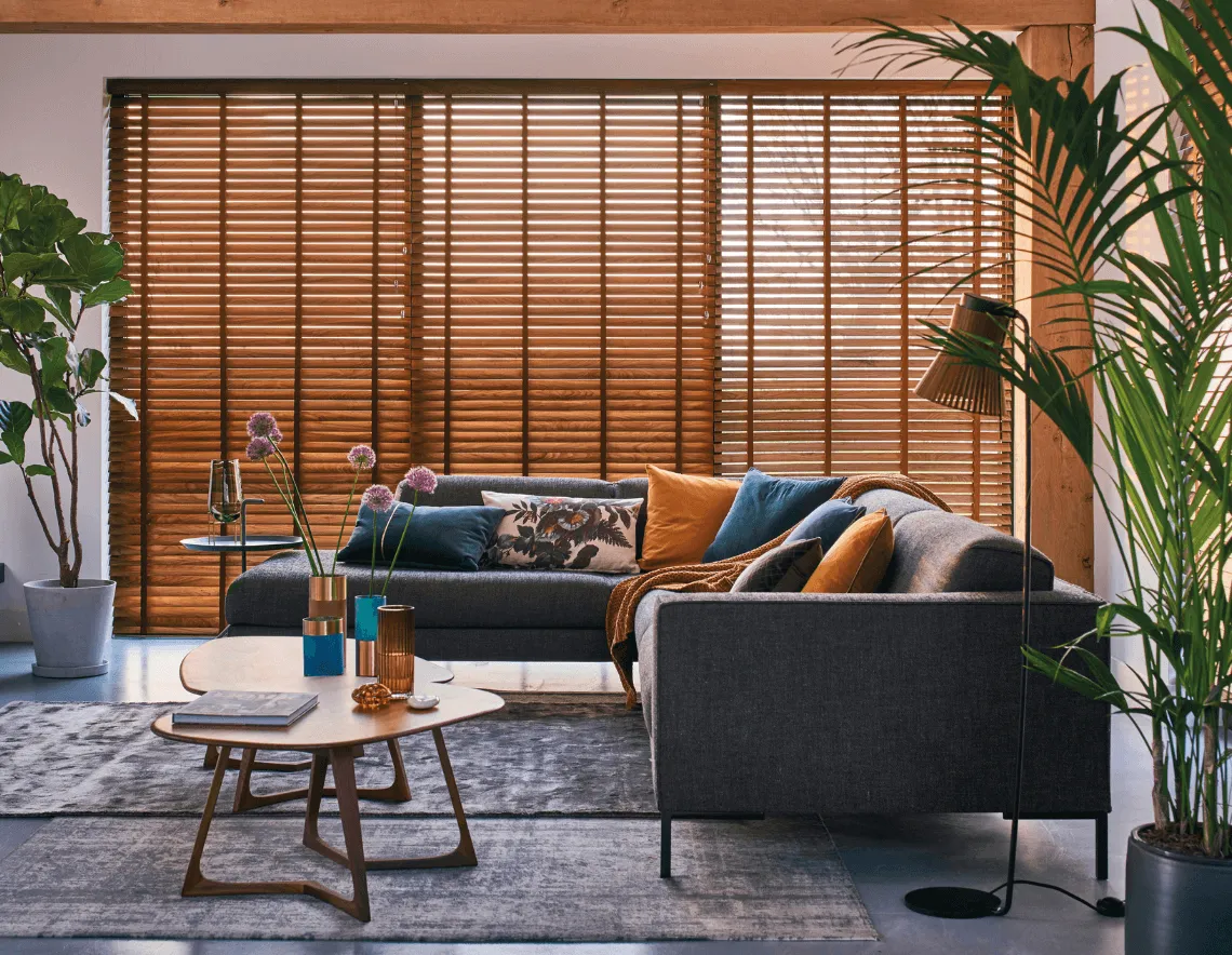 Las cortinas horizontales de madera marrón en un living con moderno sillón, plantas de interior y almohadones decorativos.